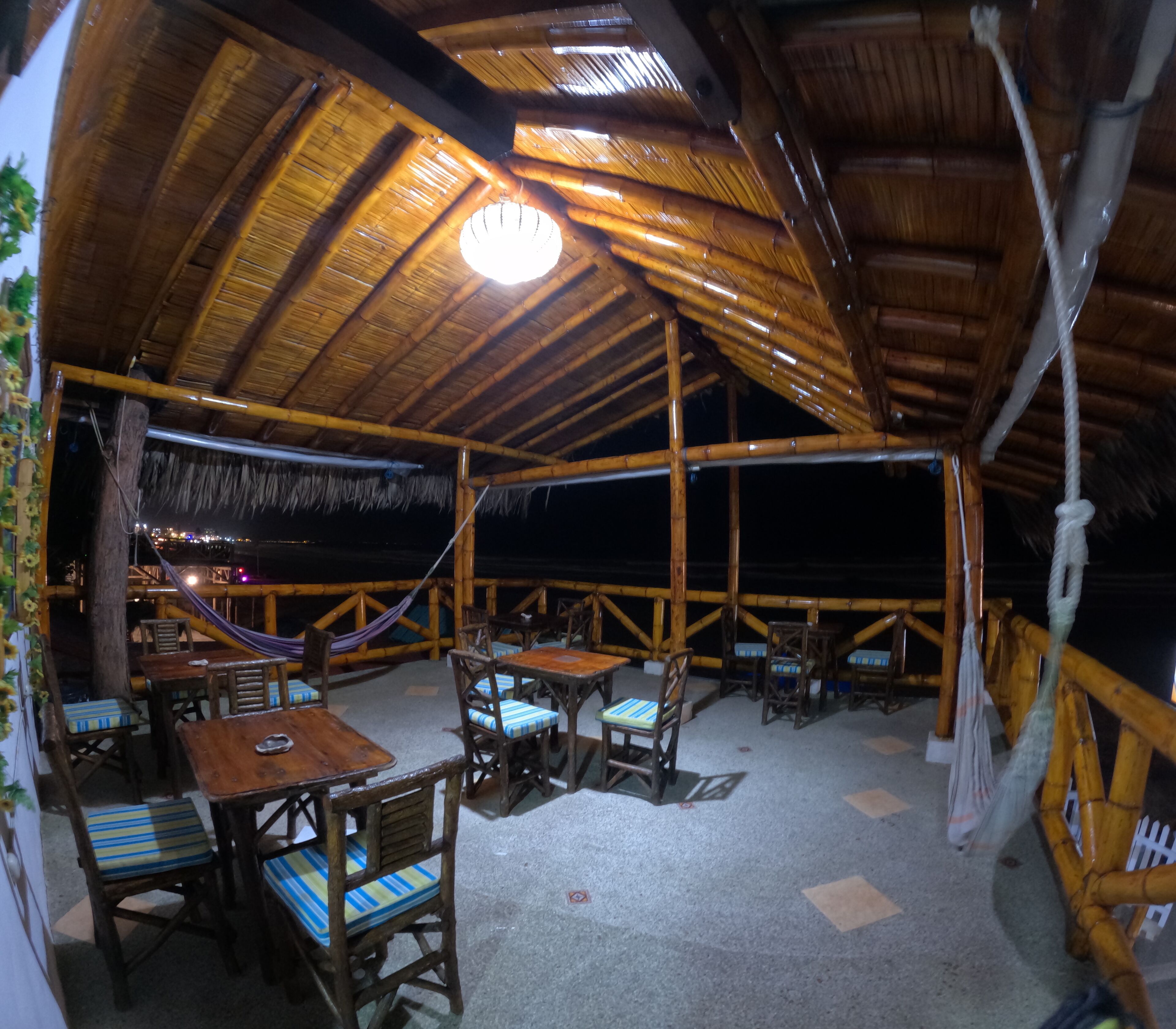 An outdoor dining area with wooden tables and chairs, a thatched roof, and a view of the night sky.