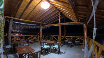 Un área de comedor al aire libre con mesas y sillas de madera, techo de paja y vistas al cielo nocturno.