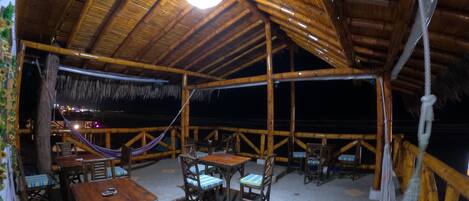 An outdoor dining area with wooden tables and chairs, a thatched roof, and a view of the night sky.