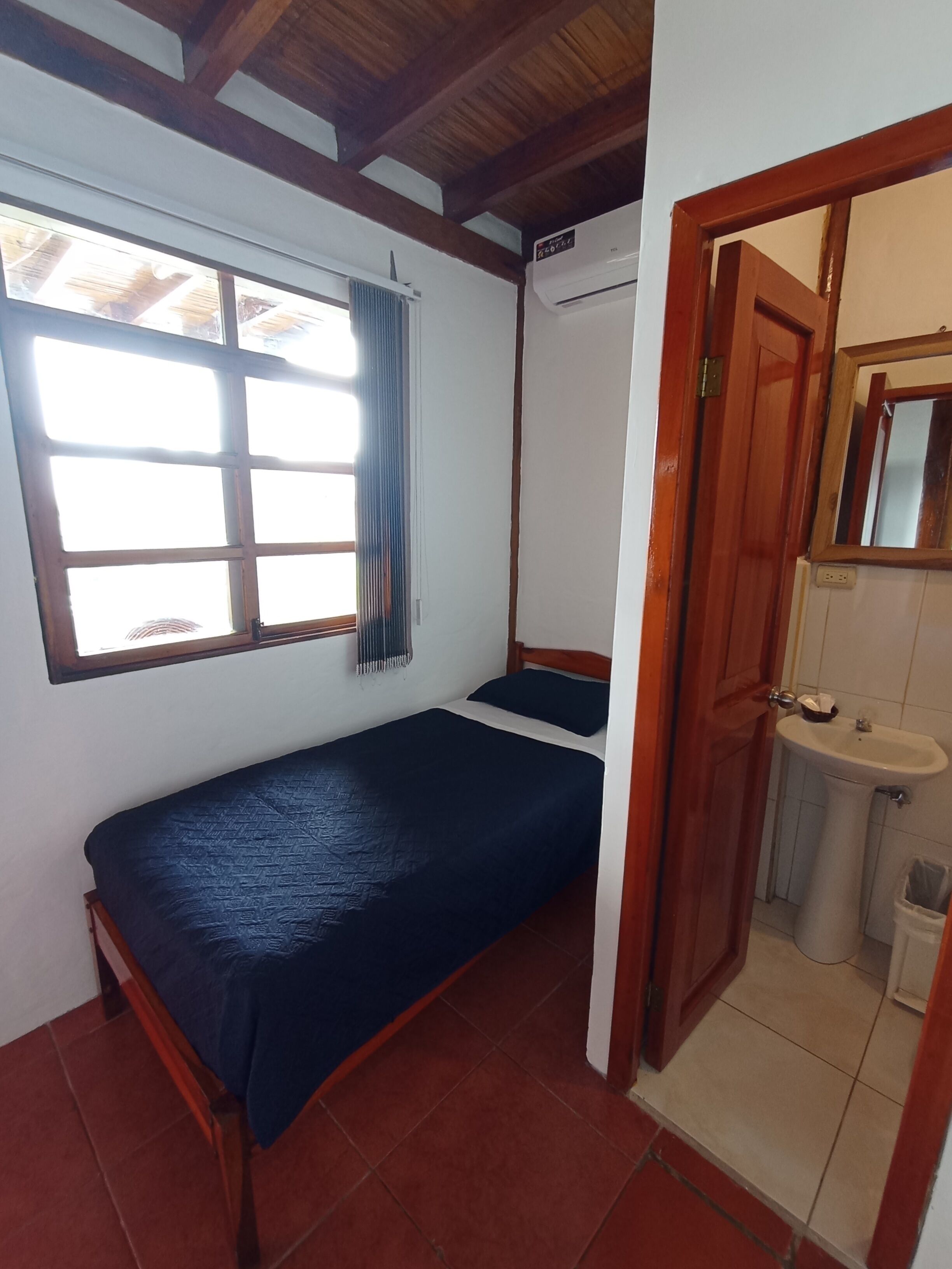 A small, compact room with a single bed, a window with curtains, a wooden ceiling, and a bathroom with a sink and mirror.