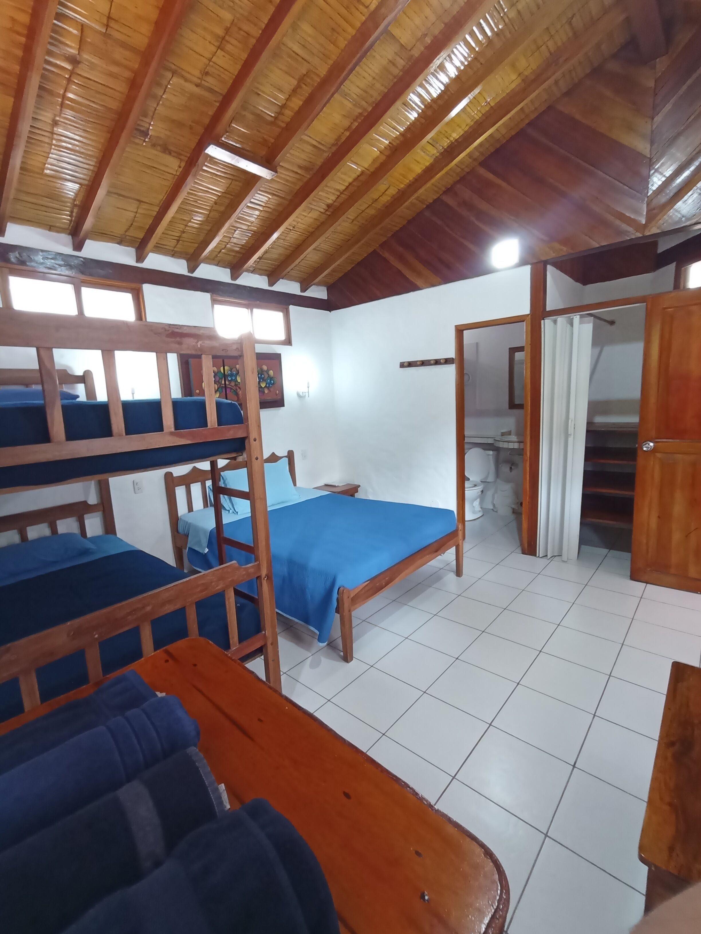 A dormitory room with bunk beds, a single bed, a wooden ceiling, and a tiled floor.