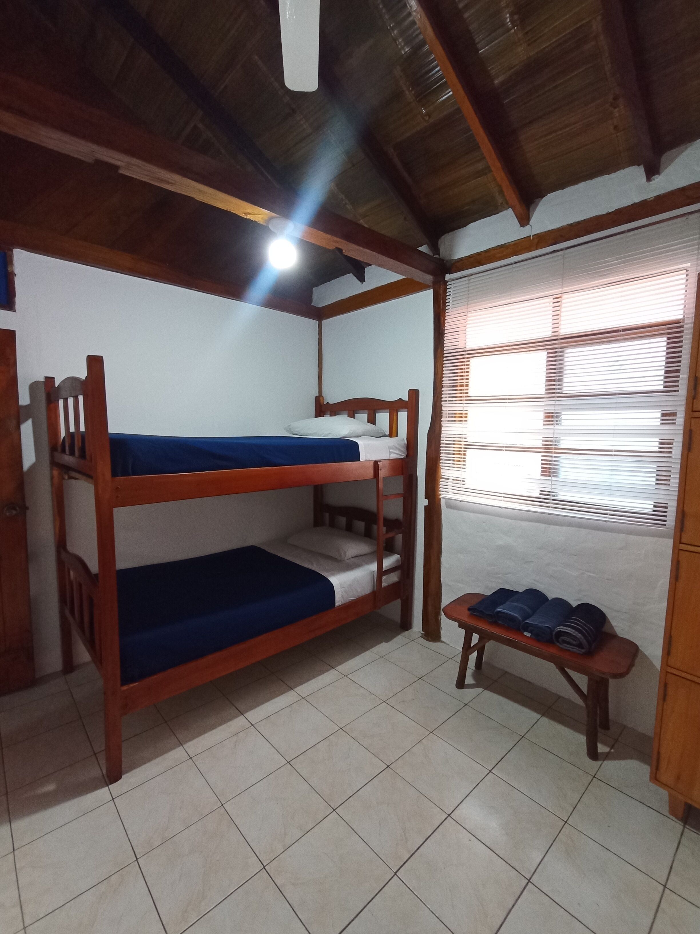 Une chambre avec des lits superposés, un plafond en bois, un banc avec des tapis enroulés et une fenêtre avec des stores.