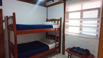 Une chambre avec des lits superposés, un plafond en bois, un banc avec des tapis enroulés et une fenêtre avec des stores.