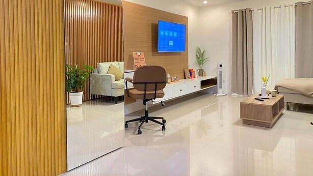 A modern living room with a flat-screen TV mounted on the wall, a wooden accent wall, a white sofa, a brown office chair, a coffee table, and a bed with curtains. - Rustic charm meets modern elegance at our wood & white retreat. (Butuan City)