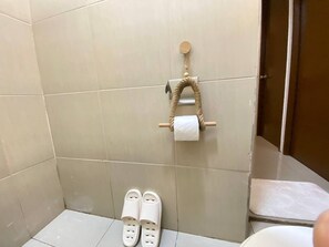 A bathroom with beige tiles, a toilet, a sink, a towel rack with a roll of paper towels, and a pair of slippers on the floor. - Rustic charm meets modern elegance at our wood & white retreat. (Butuan City)