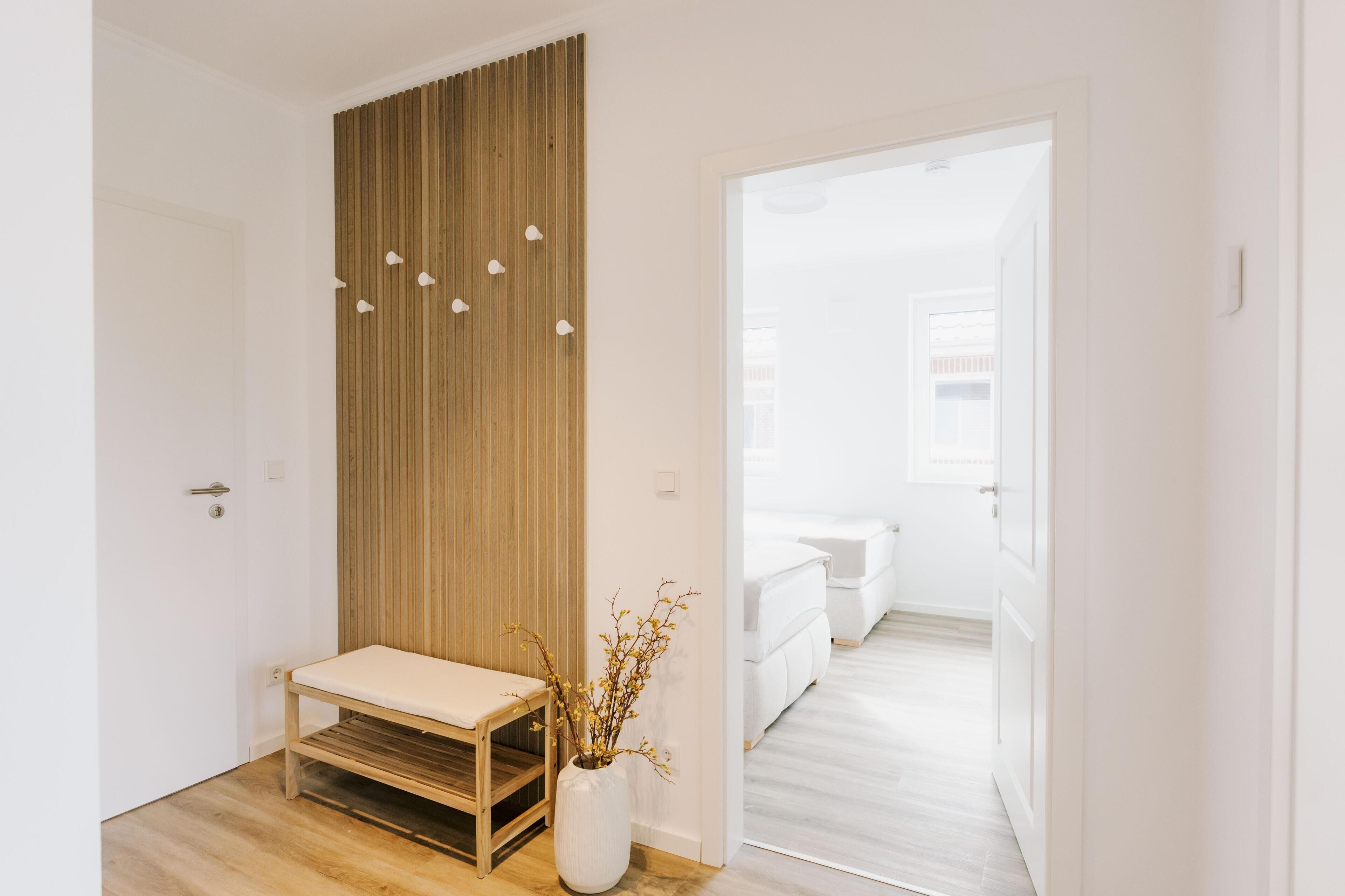 A hallway with a wooden bench, a white vase with branches, and a door leading to a bedroom.