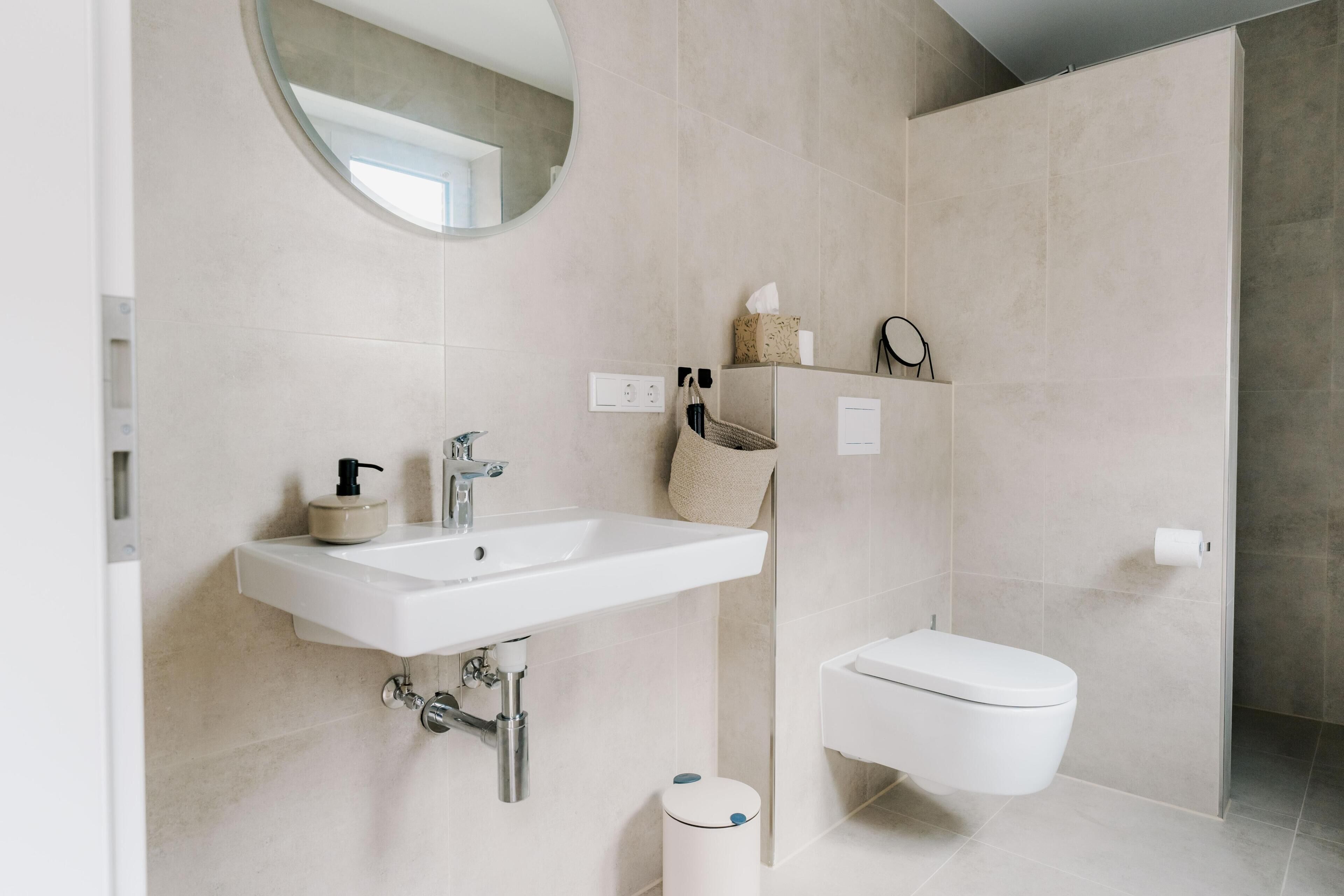 A modern bathroom with a white sink, toilet, and a round mirror.