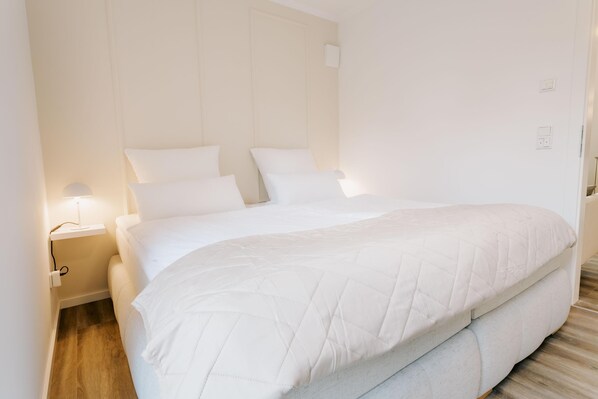 Una cama grande con edredón blanco acolchado, una mesita de noche con lámpara y suelo de madera.