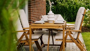 En udendørs terrasse med et træbord og stole, en murstensmur og grønne planter i baggrunden.