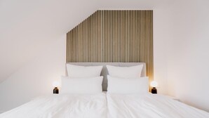A modern bedroom with a sloped ceiling, a wooden headboard, and two bedside lamps. - de See - Suite 2 Direkte Deichlage (Greetsiel)