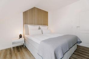 A bedroom with a bed, a bedside table, a lamp, and a wooden slat headboard. - de See - Suite 2 Direkte Deichlage (Greetsiel)