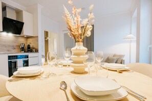 Un área de comedor con una mesa redonda preparada para cuatro personas, un jarrón con flores secas y una cocina moderna al fondo.