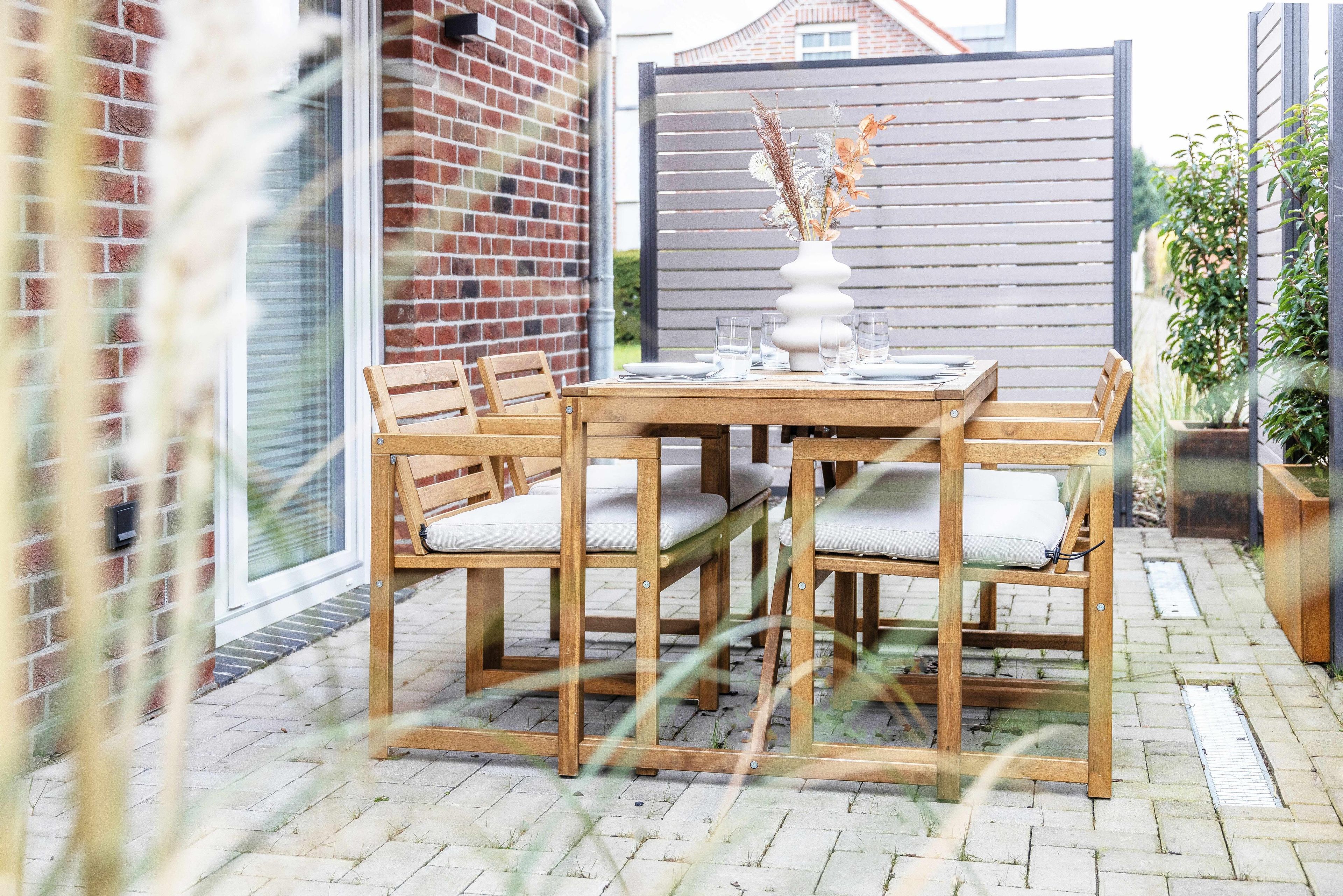 An outdoor patio with a wooden table and chairs, a white vase with dried plants, and a brick wall in the background.