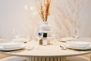 Una mesa redonda para comer con platos blancos, vasos transparentes y cubiertos. Un florero blanco con flores secas está centrado en la mesa.