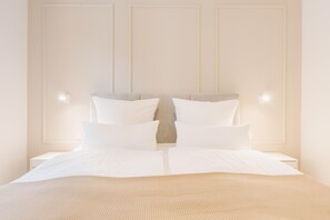 Un lit bien fait, avec du linge de lit blanc et une couverture beige à texture intéressante, flanqué de deux tables de chevet sur lesquelles sont posées des lampes.
