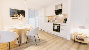 A modern kitchen with white cabinets, a built-in oven, and a dining area with a round table and chairs. - de Diek - Suite 3 Seitliche Deichlage (Greetsiel)