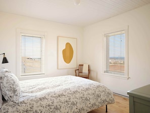 Une chambre à coucher avec un lit, deux fenêtres équipées de stores, une chaise et une œuvre d’art encadrée accrochée au mur.
