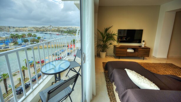 A balcony with a table and chairs, a bed with a TV and a plant, and a view of a marina and cityscape. - Luxury&stylish 2bedroom seafront, balcony GOGZR-4 (Gzira)