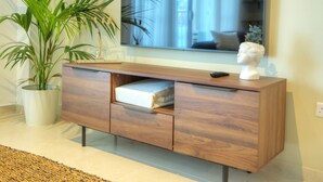 A modern living room with a large flat-screen TV mounted on the wall, a wooden cabinet with a white towel, and a large potted plant. - Luxury&stylish 2bedroom seafront, balcony GOGZR-4 (Gzira)