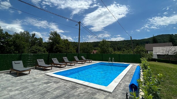 Une piscine rectangulaire, bordée d’une terrasse carrelée, entourée de chaises longues et d’une clôture verte.