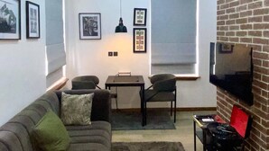 A living room with a sofa, a TV mounted on a brick wall, a table with chairs, and framed pictures on the wall. - Pearl Court Residence & Hotels (Lagos)