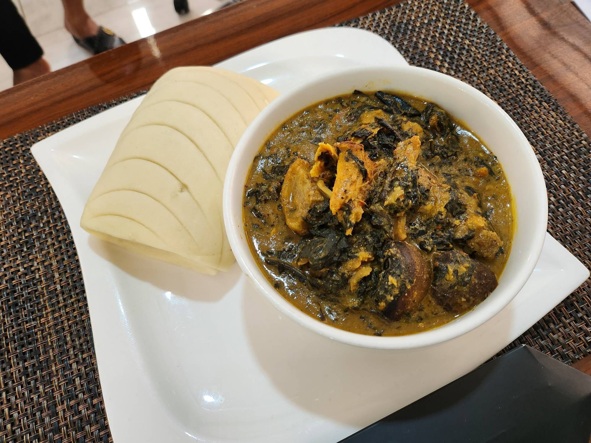 A bowl of stew with meat and greens, accompanied by slices of flatbread on a white plate.