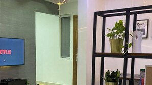 A modern hotel room with a large flat-screen TV displaying 'NETFLIX', a black metal shelf with plants, a blue sofa, and a bed with white bedding. - Pearl Court Residence & Hotels (Lagos)