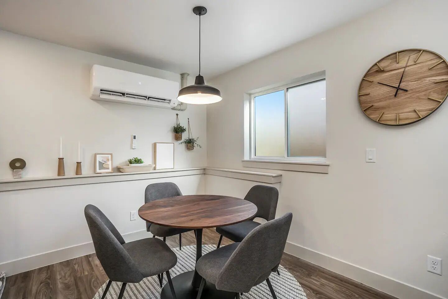 Un comedor con una mesa redonda, cuatro sillas, una lámpara colgante, un equipo de aire acondicionado, una ventana y un reloj de madera en la pared.