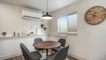 Un comedor con una mesa redonda, cuatro sillas, una lámpara colgante, un equipo de aire acondicionado, una ventana y un reloj de madera en la pared.
