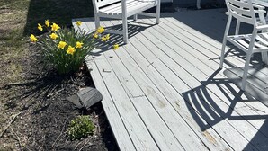 Um deck de madeira com duas cadeiras brancas, um pequeno jardim com flores amarelas e um edifício cinzento com porta amarela.