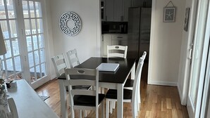 Uma área de refeições com mesa e cadeiras, uma cozinha ao fundo e vista através de grandes janelas.