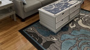 Uma sala de estar com um sofá modular cinza, uma mesa de centro branca com design de bicicleta, um tapete estampado e uma janela com persianas.