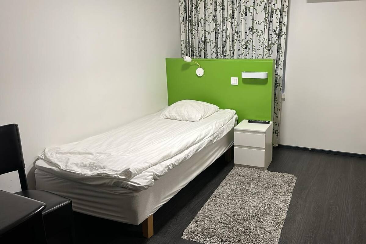 A small, neatly arranged bedroom with a single bed, a bedside table, a chair, and a green headboard.