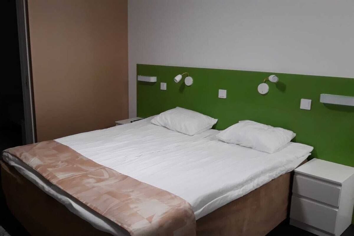 A neatly made bed with white linens and a brown accent throw, a green headboard, and a white nightstand with a drawer.