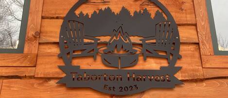 A wooden cabin with a carved logo featuring a campfire, two chairs, and trees, and the text "Taborton Harvest Est 2023".