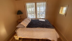 A bedroom with a bed, a nightstand, a lamp, a window with a view of trees and snow, and wooden walls and floor. - Taborton Harvest Timber Trails
Unique cabin experience (Sand Lake)