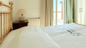 Ein Hotelzimmer mit Bett, Nachttisch mit Lampe, Fenster mit Vorhängen und Blick ins Freie.
