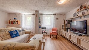 A cozy living room with a sofa, armchair, and a television. There are bookshelves and a decorative shelf with items.