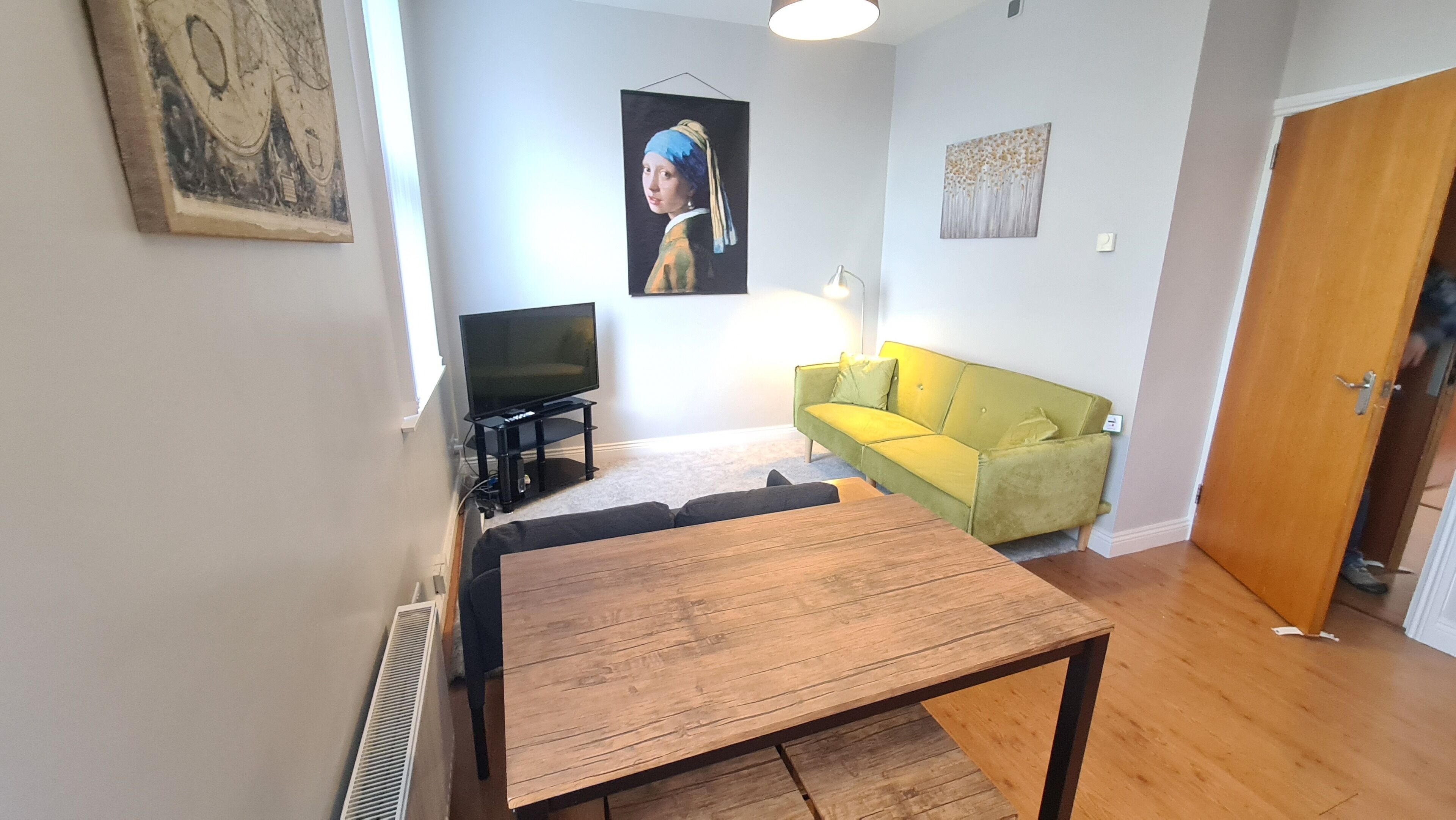A living room with a wooden table, a green sofa, a TV, and artwork on the walls.