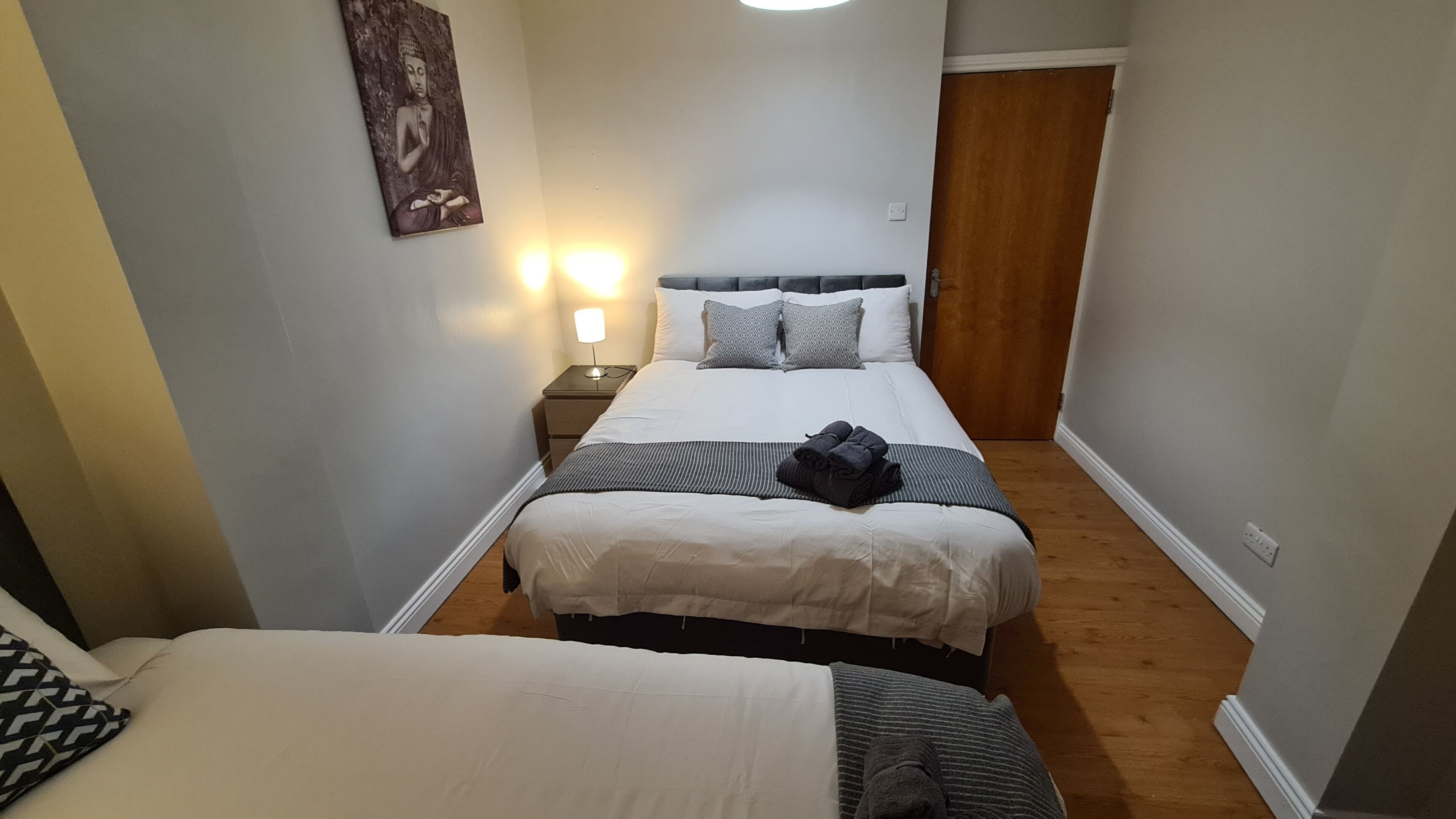 A narrow hotel room with two beds, a nightstand with a lamp, and a wall-mounted artwork.