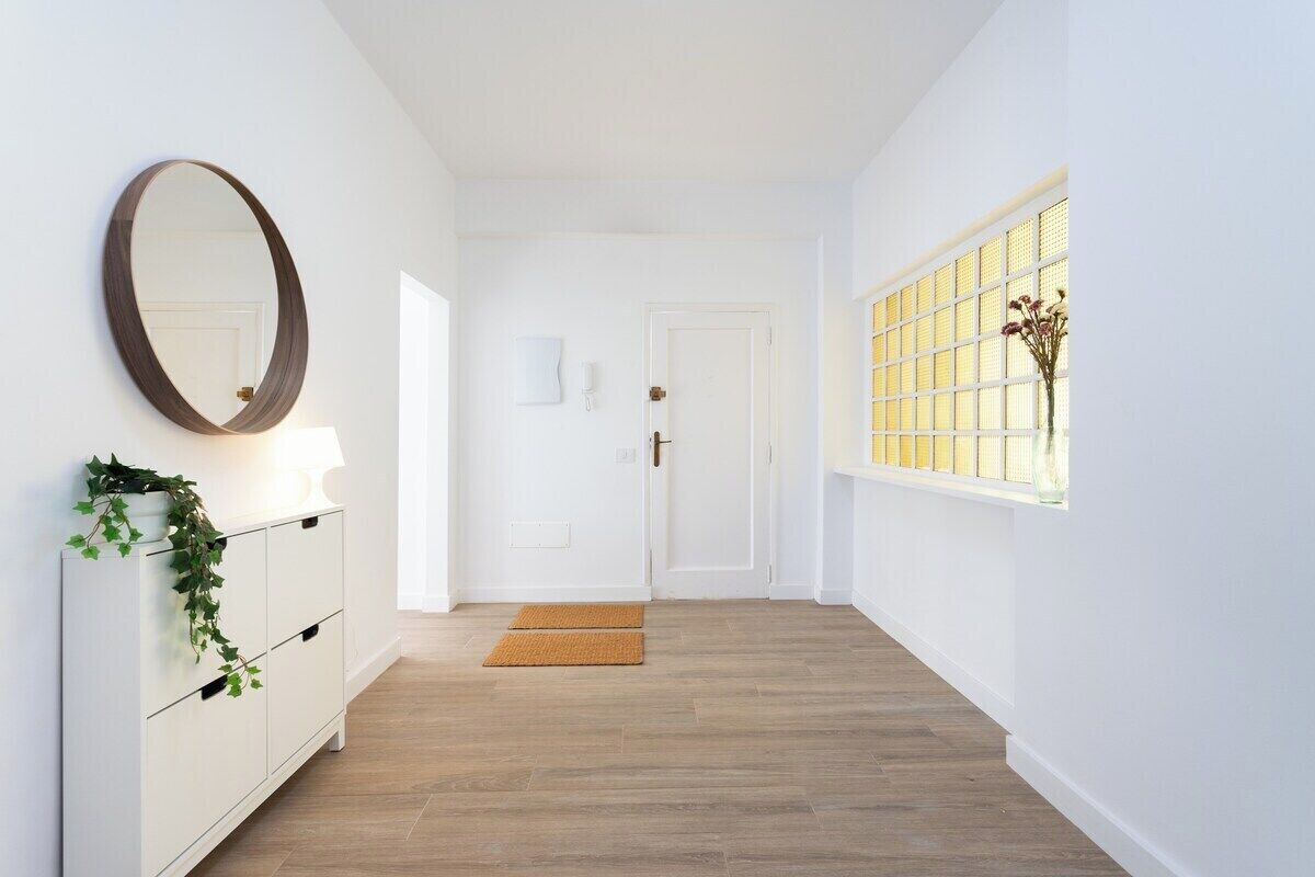 Un couloir avec une porte blanche, un miroir, un meuble blanc orné d’une plante et une fenêtre donnant sur un paysage.