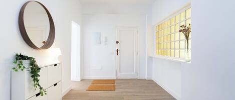 Un couloir avec une porte blanche, un miroir, un meuble blanc orné d’une plante et une fenêtre donnant sur un paysage.