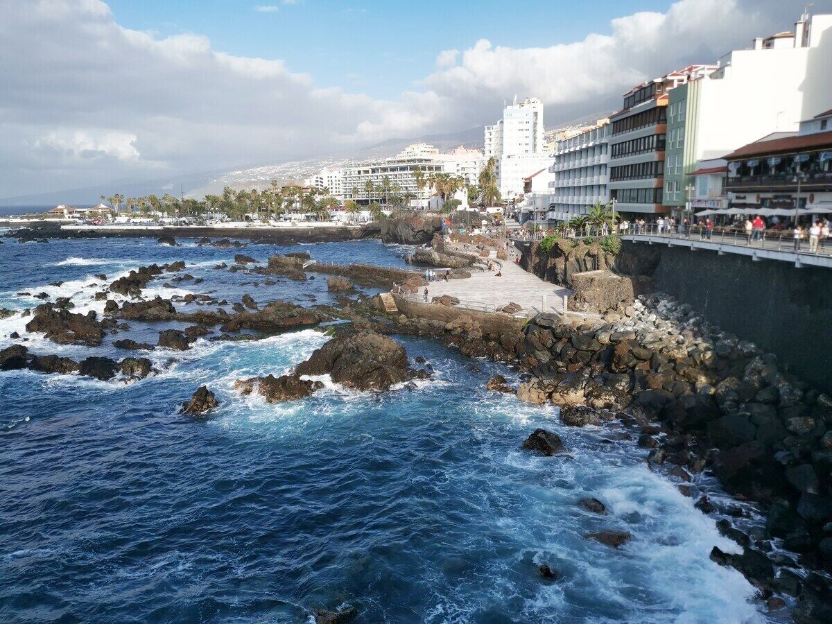 Vista costanera amb aigües rocoses, un passeig marítim i edificis al fons.