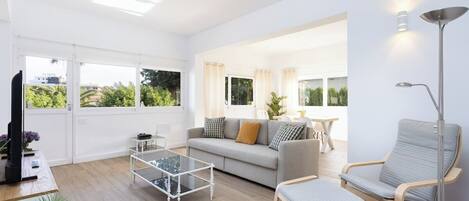 Amplia sala de estar con sofá, dos sillones, una mesa de centro y televisor. Ventanas grandes ofrecen vista a la vegetación exterior.