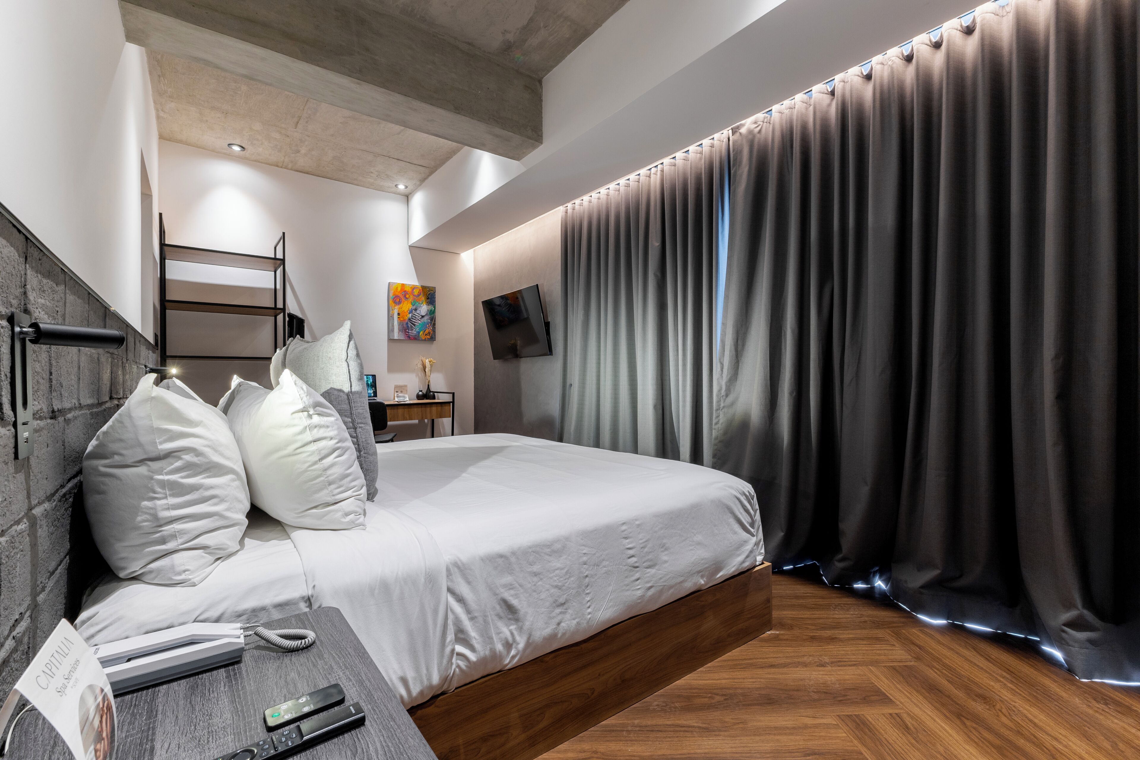 Una camera d'albergo moderna con un letto grande, una testiera in legno, un comodino con telefono e telecomando, una scrivania piccola con una lampada e un'ampia finestra con tende.