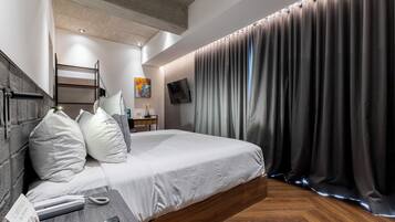 Una camera d'albergo moderna con un letto grande, una testiera in legno, un comodino con telefono e telecomando, una scrivania piccola con una lampada e un'ampia finestra con tende.