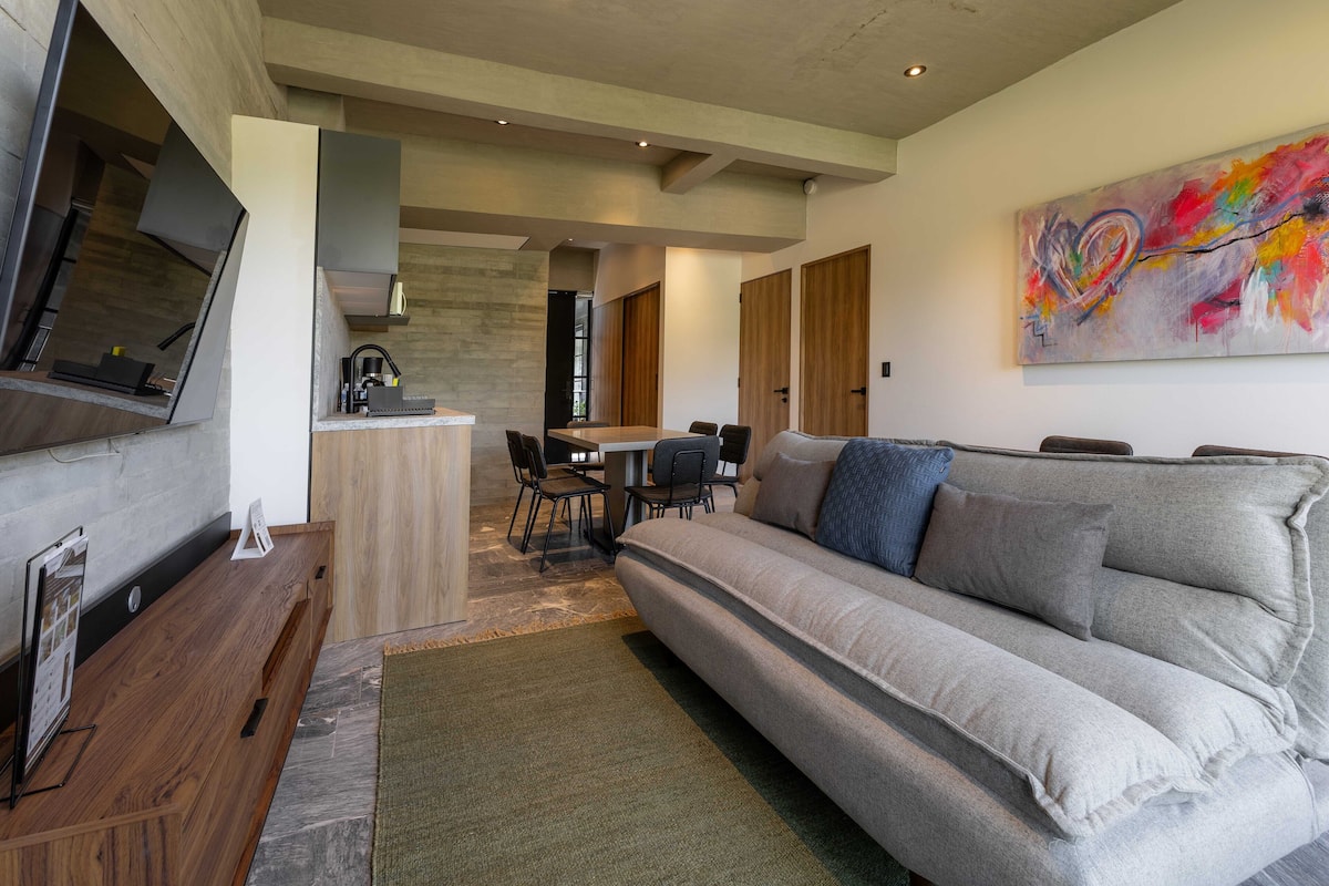 Una sala de estar moderna con un sofá gris, una mesa de centro de madera y un televisor montado en la pared. Se observa una zona de cocina al fondo, con una mesa de comedor y sillas.