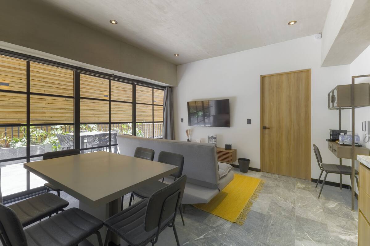Una sala de estar moderna con una mesa de comedor, sofá y sillas. Hay un televisor montado en la pared, una puerta de madera y una ventana con persianas.