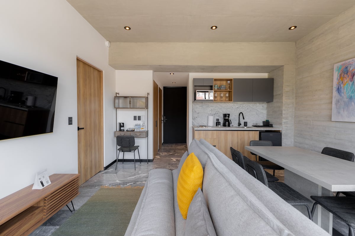 Una cocina moderna con electrodomésticos integrados, un comedor con sofá gris y cojín amarillo, y un televisor montado en la pared.