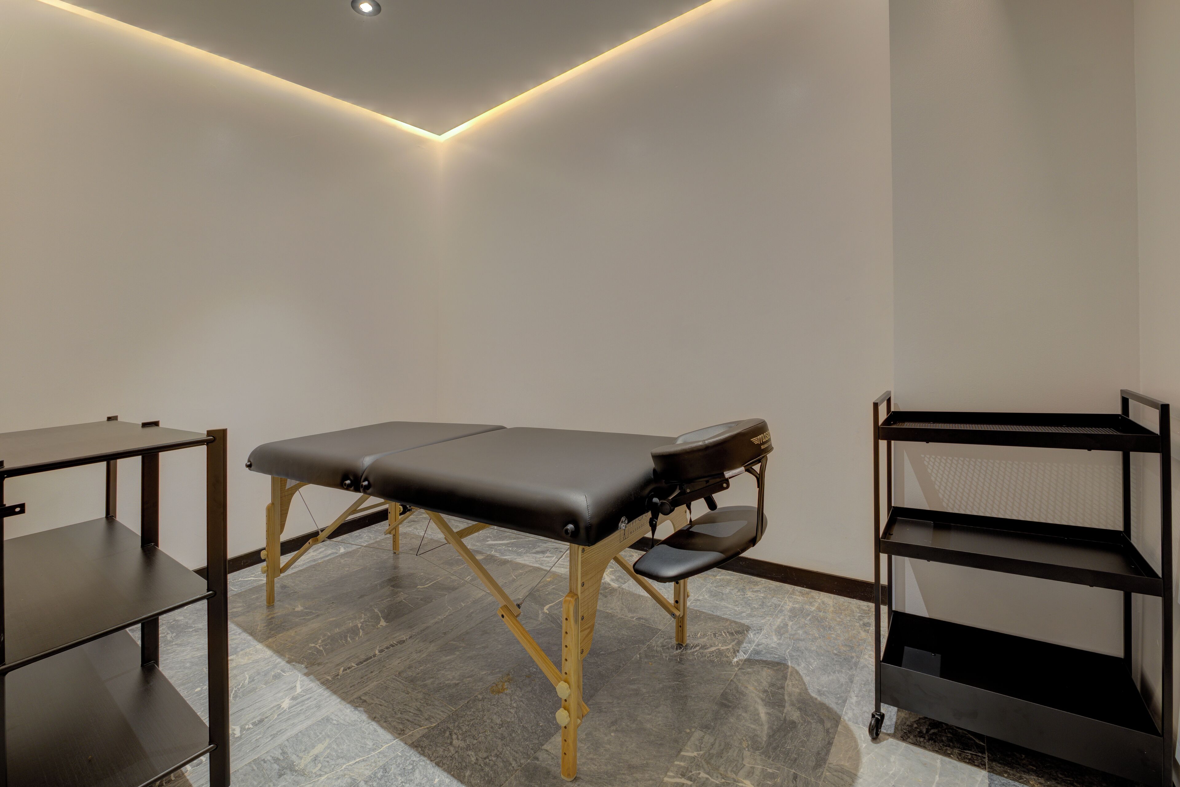Una sala dedicata ai trattamenti con lettino da massaggio, pavimento in legno e scaffalunità in metallo nero.